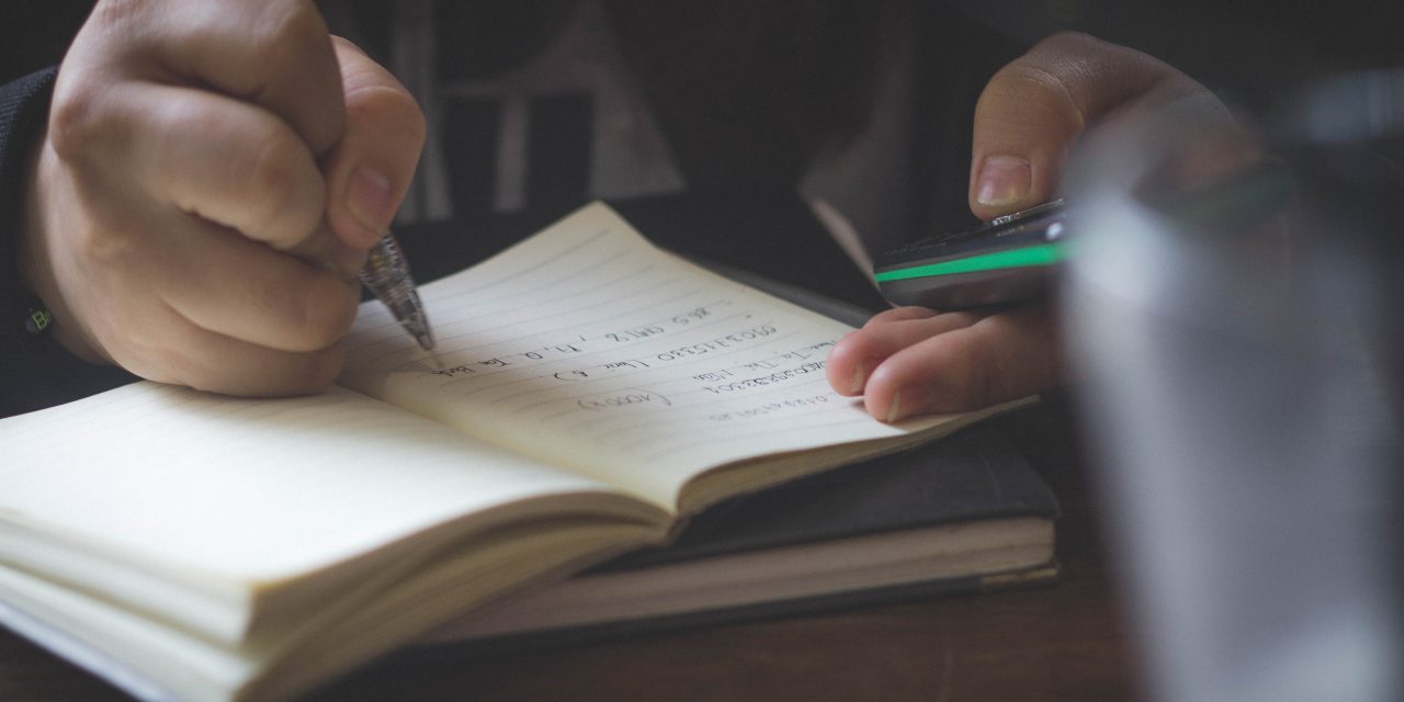 Close up of man on his smartphone while writing in a notebook.